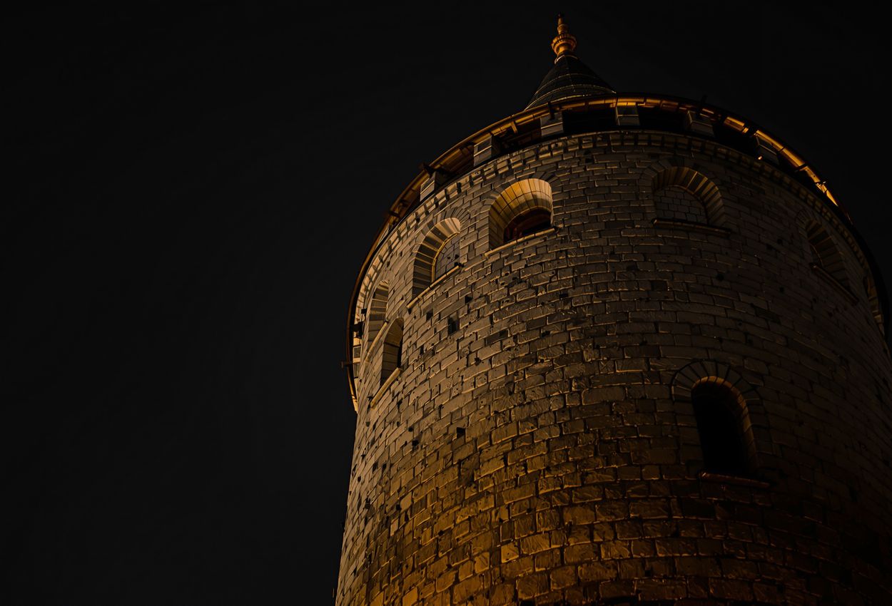 Close‑up nighttime image of the historic Daliborka Tower in Prague Castle, seen from a low angle with dramatic spotlight lighting its textured stone walls, light trails in the air, and the dark sky above, evoking the medieval prison’s history.